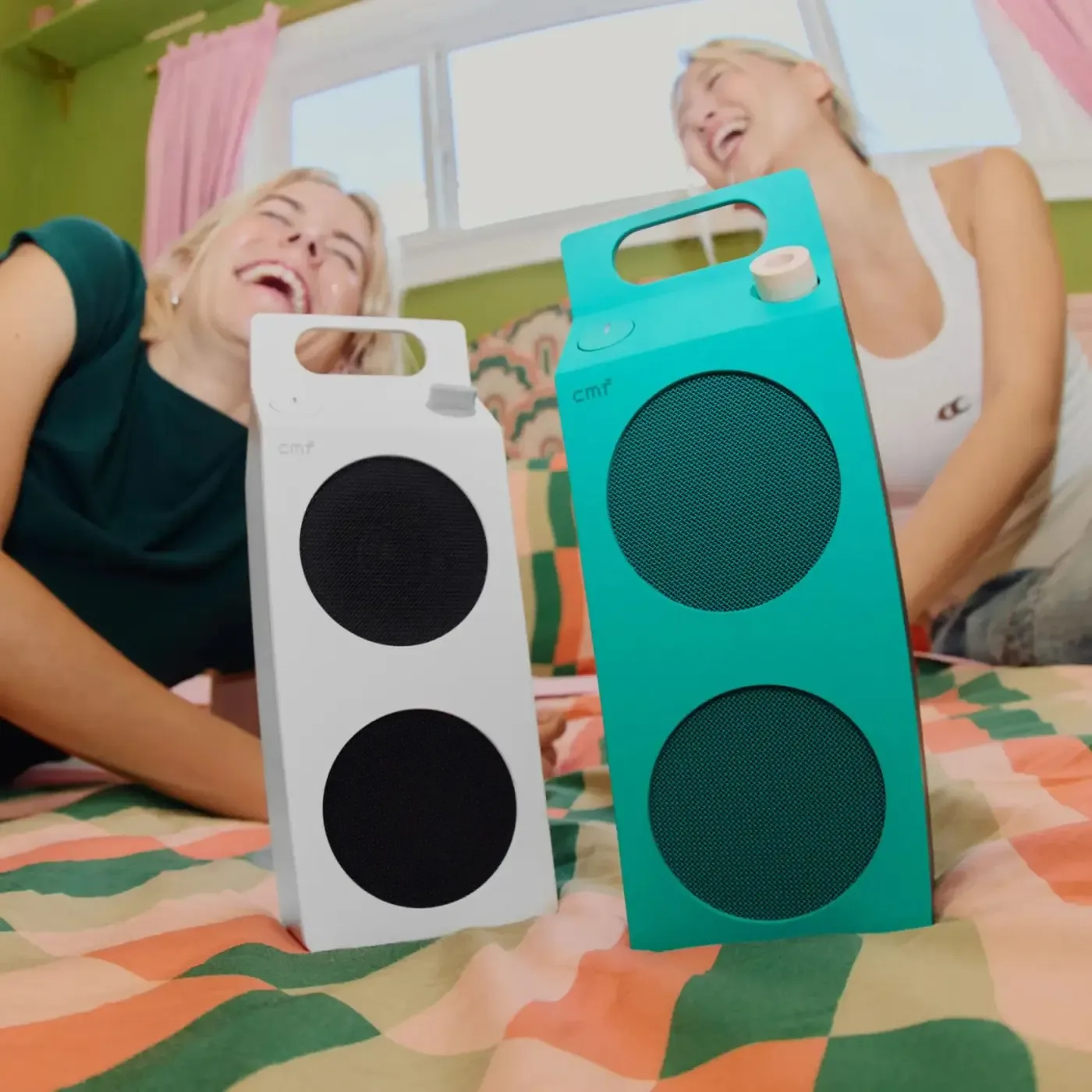 Two women laughing on a bed with white and turquoise Speaker M units in front
