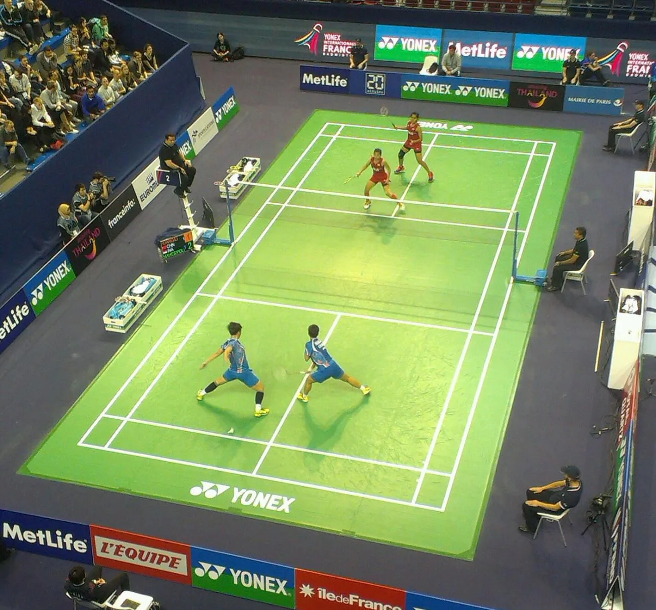 A green badminton court at the Internationaux de France (French Open) seen from above, with doubles players and spectators around it